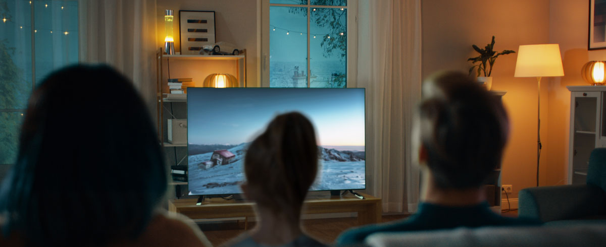 Eine Familie sitzt zusammen im Wohnzimmer und schaut gemeinsam auf einem großen Fernseher fern